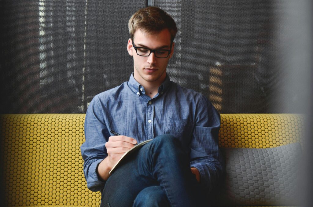 A young man in glasses writes in a notebook while sitting on a stylish couch indoors.