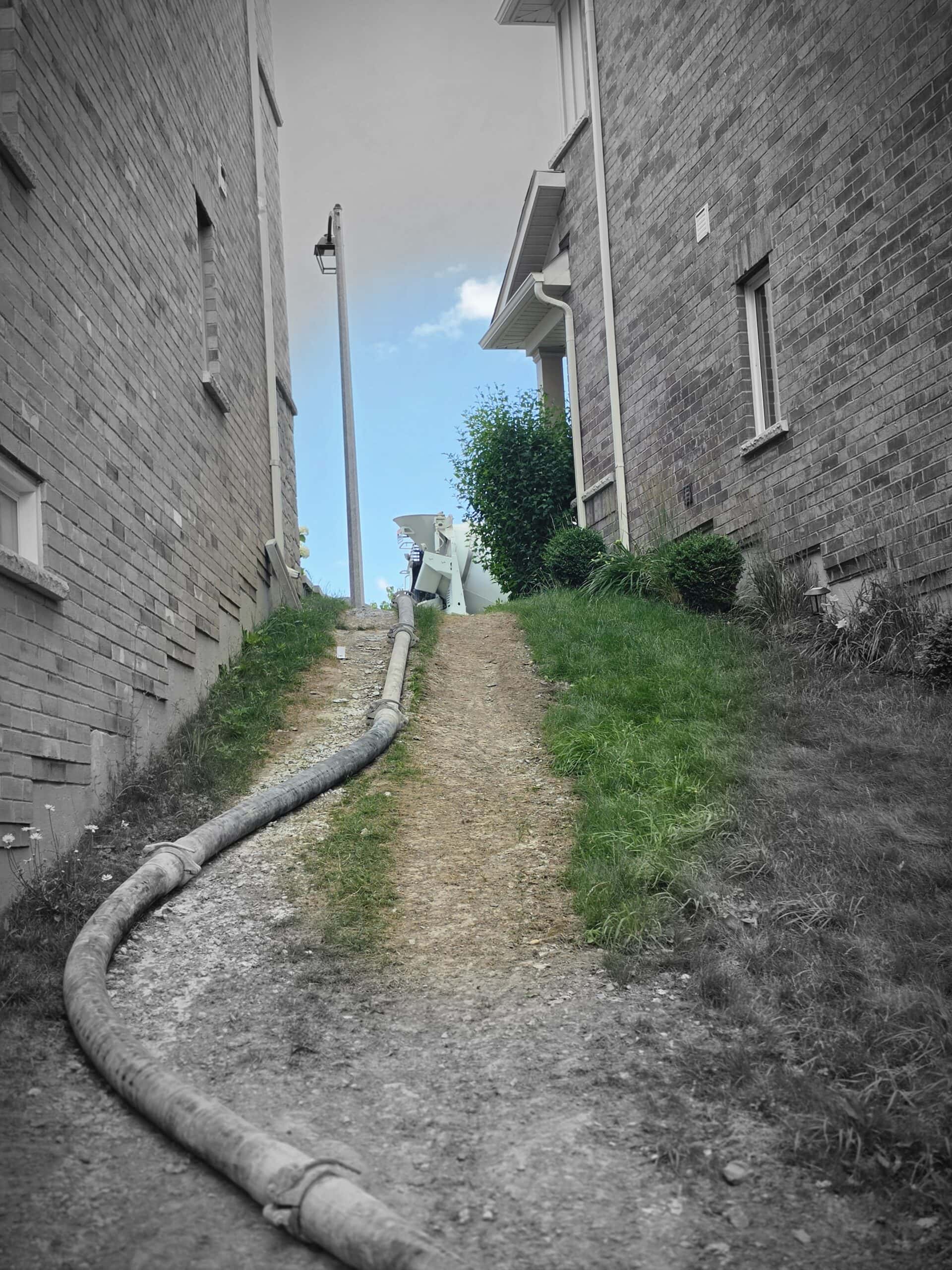 Concrete pump hose routed through a narrow side yard to reach a hard-access pour area.